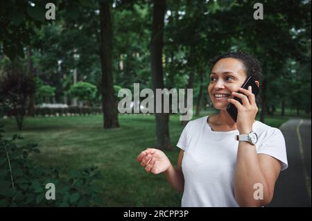 Charmante, multiethnische junge Frau in einem weißen T-Shirt, lächelnd, telefonisch, während sie an einem Sommertag durch die Gasse eines Stadtparks schlendert. Menschen. Stockfoto