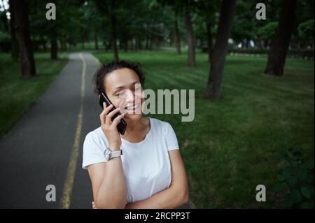 Schöne Touristin, junge Frau in weißem T-Shirt, lächelnd, telefoniert mit dem Handy, während sie an einem Sommertag durch die Gasse eines Stadtparks schlendert. Peo Stockfoto