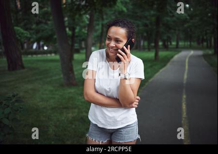 Wunderschöne, multiethnische junge Frau in weißem T-Shirt, lächelt, telefoniert mit dem Handy, während sie an einem Sommertag die Gasse eines Stadtparks entlanggeht. Peo Stockfoto