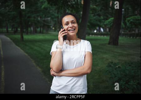 Porträt einer schönen jungen Frau, die lächelt, mit dem Handy spricht, während sie an einem Sommertag durch die Gasse eines Stadtparks schlendert. Menschen. Kommunikation. Stockfoto