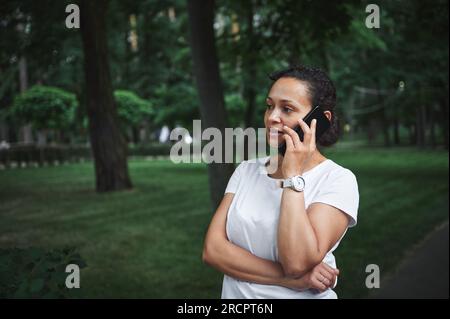 Selbstbewusste, multiethnische junge, hübsche Frau in einem weißen T-Shirt, lächelnd, telefoniert am Sommertag, während sie durch die Gasse eines Stadtparks schlendert. Peo Stockfoto