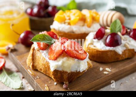 Beeren Toast Frühstück, gesundes Essen. Sandwich mit Kirsche, Erdbeeren, Weichkäse und Honig auf Holzbrett auf Steinhintergrund. Stockfoto