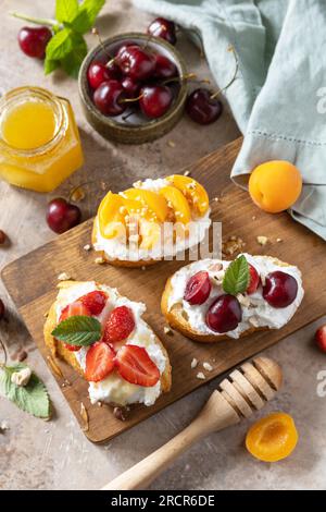 Sandwich mit Kirsche, Erdbeeren, Weichkäse und Honig auf Holzbrett auf Steinhintergrund. Beeren Toast Frühstück, gesundes Essen. Stockfoto