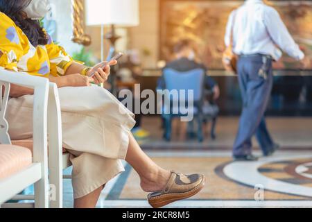 Die Hände der weiblichen Reisenden tragen eine Gesichtsmaske und sitzen auf dem Smartphone, während sie auf den Hoteleincheck in der Hotellobby warten. Eine Geschäftsfrau, die in der Hotellobby wartet Stockfoto