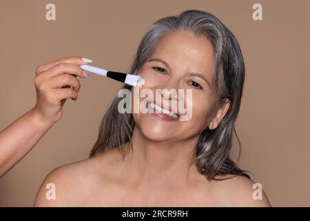 Fröhliche ältere weiße Dame genießt Anti-Aging-Behandlung, Kosmetikerin, trägt Creme Maske mit Bürste Stockfoto