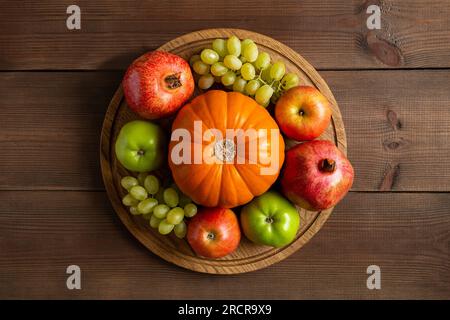 Im Herbst gibt es noch reife Früchte, orangefarbener Kürbis, Äpfel, Trauben und Granat. Herbsternte auf rundem Holzschneidbrett und brauner, flacher Unterlage Stockfoto