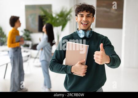 Aufgeregter europäischer männlicher Student, der Daumen hoch zeigt und lächelt und Bildungskurse empfiehlt Stockfoto