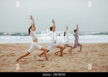 Sand, Brandung und Ruhe. Junge Leute Yoga-Kursteilnehmer stehen in Warrior One Pose, trainieren am Strand Stockfoto