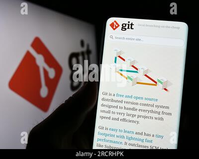 Person, die ein Mobiltelefon mit einer Webseite des verteilten Versionskontrollsystems Git auf dem Bildschirm vor dem Logo hält. Konzentrieren Sie sich auf die Mitte des Telefondisplays. Stockfoto