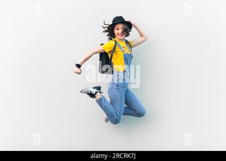 Portrait einer attraktiven, überfreulichen Hipsterin in blauem Jeansanzug, gelbem T-Shirt und schwarzem Hut, springen, gute Laune haben, Glück ausdrücken. Studioaufnahmen im Innenbereich isoliert auf grauem Hintergrund. Stockfoto