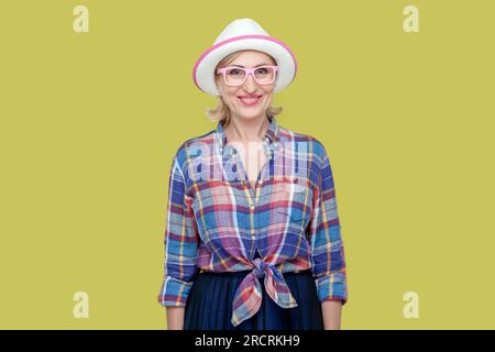 Portrait einer lächelnden, freudigen Seniorin mit kariertem Hemd, Hut und Brille, die mit einem zahnigen Lächeln in die Kamera schaut und positive Emotionen ausdrückt. Innenstudio-Aufnahme isoliert auf gelbem Hintergrund Stockfoto