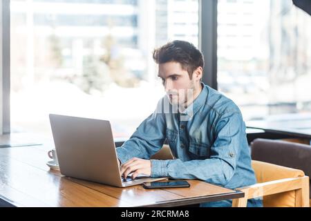 Porträt eines hochkonzentrierten jungen Freiberuflers in Blue Jeans Hemd, der am Laptop arbeitet, auf der Tastatur tippt, mit Projekt arbeitet. Innenaufnahmen in der Nähe eines großen Fensters, Café-Hintergrund. Stockfoto