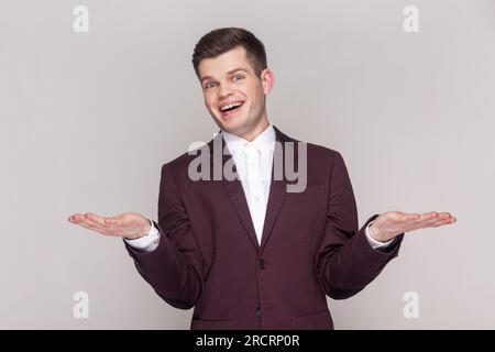 Das Porträt eines fröhlichen, positiven, optimistischen, gutaussehenden Mannes verteilt die Hände, lädt Gäste ein, zeigt Kopien auf Handflächen, trägt violetten Anzug und weißes Hemd. Aufnahme im Studio isoliert auf grauem Hintergrund. Stockfoto