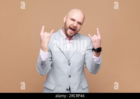 Das Porträt des kahlbärtigen Mannes Rocker genießt positive Vibes, hört Rock 'n' Roll, coole Musik im Club, zeigt Horngesten, trägt graue Jacke. Studioaufnahmen isoliert auf braunem Hintergrund. Stockfoto