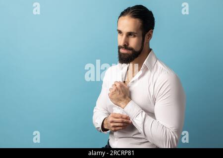 Porträt eines gutaussehenden, selbstbewussten, nachdenklichen Mannes mit Bart, der ein weißes Hemd trägt und Manschettenknöpfe verdeckt, auf einem wichtigen Event. Innenstudio, isoliert auf blauem Hintergrund. Stockfoto