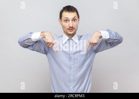 Porträt eines ernsthaften, strengen, jungen Erwachsenen, der mit beiden Zeigefingern nach unten zeigt und sagt, hier und jetzt, in einem hellblauen Hemd. Studioaufnahmen im Innenbereich isoliert auf grauem Hintergrund. Stockfoto
