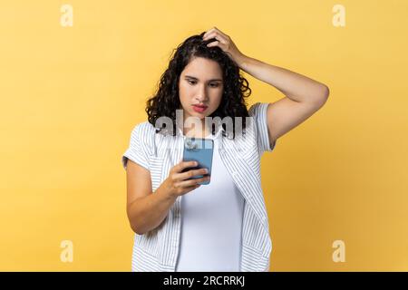 Porträt einer pensiven, wunderschönen, jungen erwachsenen Frau mit dunklem, welligem Haar, die ein Handy benutzt und denkt, was sie beantworten soll, hält die Hand am Kopf. Innenstudio-Aufnahme isoliert auf gelbem Hintergrund. Stockfoto
