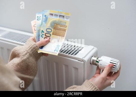 Eine Frau, die Geld vor dem Heizkörper hält. Bezahlung für Heizung im Winter Stockfoto