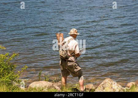 Ein älterer Mann über 70 steht neben einem See und fischt mit einer Kniestütze und einem Rucksack Stockfoto