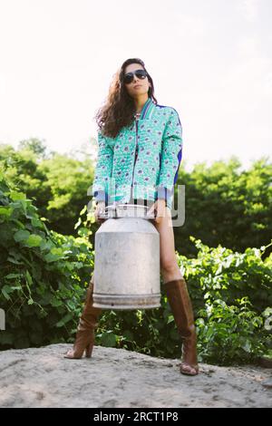 Junge, modische Bauernfrau, die vor der Kamera posiert, während sie die Milchdose hält Stockfoto