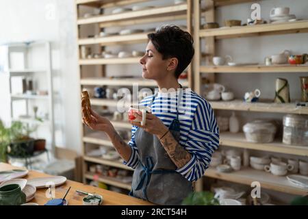 Eine junge Frau in der Töpferei trinkt Tee aus einer handgemachten Tasse, Kleinunternehmen, Keramikbesitzer-Konzept Stockfoto