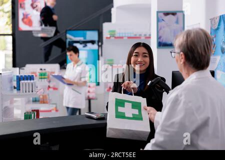 Älterer Apotheker, der dem lächelnden Käufer an der Apothekenpreiskasse eine Medikamententüte gibt. Zufriedener Kunde, der eine Bestellung von einem Pharmazeutikmitarbeiter an der Rezeption erhält Stockfoto