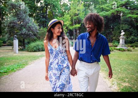 Ein multiethnisches Paar spaziert im Sommer in einem öffentlichen Park und hält Händchen Stockfoto