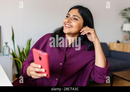Glückliche, entspannte junge indische Frau, die zu Hause Handy benutzt Stockfoto