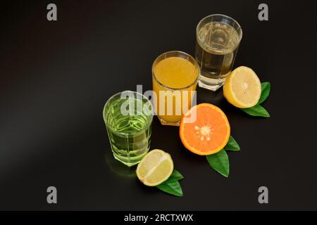 Drei große Gläser mit verschiedenen Zitrussäften auf schwarzem Hintergrund, neben den Hälften von reifer Orange, Zitrone und Limette mit Blättern. Nahaufnahme. Stockfoto