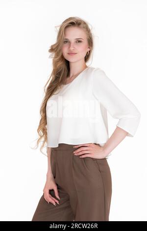 Porträt einer eleganten blonden Frau mit langen Haaren, die in die Kamera schaut. Wunderschöne junge Erwachsene Frau in weißer Bluse und brauner Hose Stockfoto