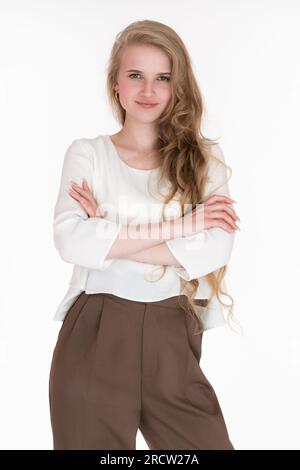 Lächelnde junge blonde Frau mit gekreuzten Armen, die in die Kamera schaut. Herrisch-junge Geschäftsfrau, 21 Jahre alt, in weißer Bluse und brauner Hose Stockfoto