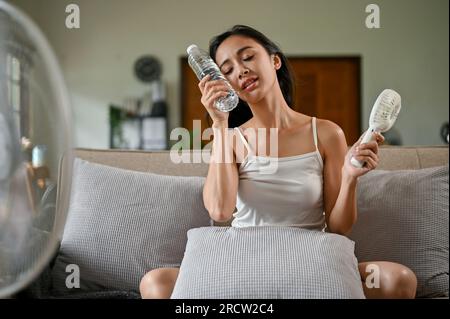 Eine müde und heiße asiatische Frau versucht sich an einem heißen Sommertag zu Hause zu kühlen, indem sie vor einem elektrischen Ventilator sitzt und ihr Gesicht verpustet Stockfoto