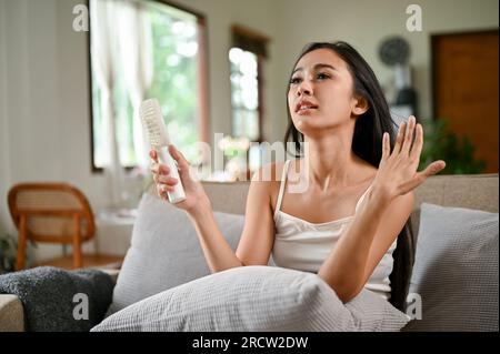 Eine müde und überhitzte asiatische Frau, die sich durch einen Hitzeanfall unwohl fühlt, verwendet einen elektrischen Ventilator, um sich abzukühlen, während sie sich auf einer Couch ausruht Stockfoto