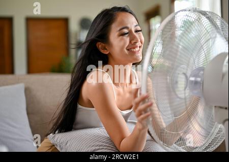 Eine glückliche und hübsche asiatische Frau atmet frische Luft aus einem elektrischen Ventilator, während sie sich an einem heißen Sommertag auf einer Couch in ihrem Wohnzimmer ausruht. Stockfoto