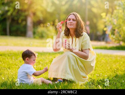 Eine Mutter spielt mit einem Baby in der Natur und bläst Seifenblasen. Glückliches Kind und mutter spielen auf dem grünen Gras im Sommerpark. Der Junge ist ungefähr zwei Ye alt Stockfoto
