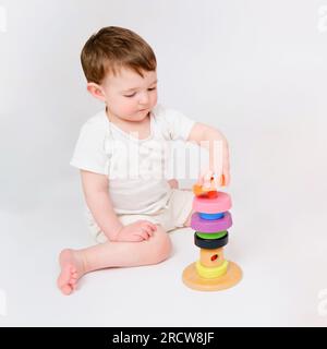 Fröhliches Baby spielt pädagogische Spielzeuge auf weißem Studiohintergrund. Porträt eines lächelnden Kindes, das auf dem Boden sitzt. Ein Kind, das etwa zwei Jahre alt ist ( Stockfoto