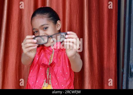 Ein indisches kleines hübsches Mädchen, das mit ihrer Brille in verschiedenen Posen in rotem Kleid rumfummelt. Stockfoto