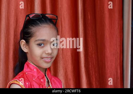 Ein indisches kleines hübsches Mädchen, das mit ihrer Brille in verschiedenen Posen in rotem Kleid rumfummelt. Stockfoto