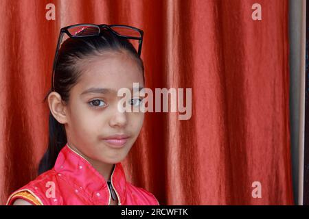 Ein indisches kleines hübsches Mädchen, das mit ihrer Brille in verschiedenen Posen in rotem Kleid rumfummelt. Stockfoto