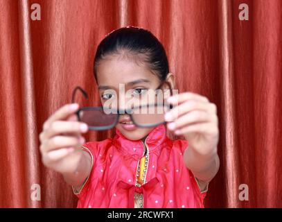 Ein indisches kleines hübsches Mädchen, das mit ihrer Brille in verschiedenen Posen in rotem Kleid rumfummelt. Stockfoto