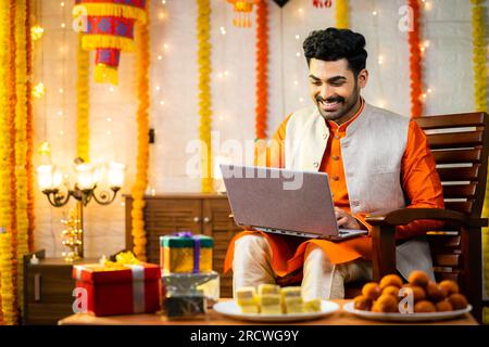 Glücklicher junger indischer Mann, der während der diwali Festival Feier im dekorierten Haus ein Notebook benutzte, vor Geschenken und Geschenken, indem er in die Kamera schaute - Konzept Stockfoto
