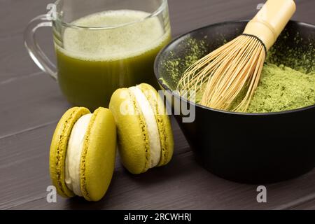 Bambus-Schneebesen in einer Schüssel mit Matcha-Pulver. Zwei Makkaroni-Kuchen und eine Tasse Matcha-grünen Tee. Schließen. Dunkler Holzhintergrund Stockfoto