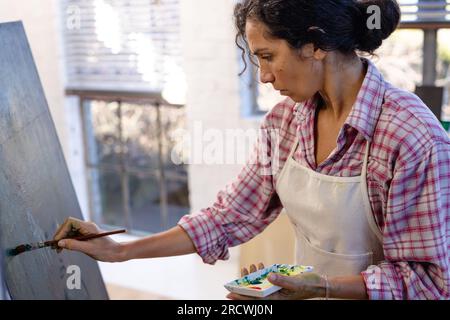 Birassische Frau, die Schürzenmalerei auf Staffelei trägt, mit Pinsel im Heimstudio Stockfoto