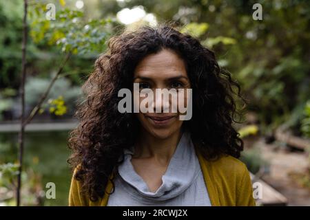 Porträt einer glücklichen, birassischen Frau mit dunklen, lockigen Haaren, die im Garten lächeln Stockfoto