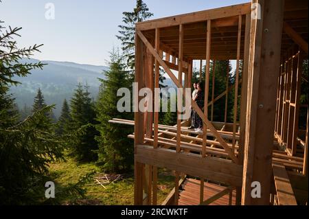 Mann und Frau inspizieren ihr zukünftiges Holzrahmenhaus, eingebettet in den Bergen in der Nähe des Waldes. Ein junges Paar auf der Baustelle am frühen Morgen. Konzept des zeitgenössischen ökologischen Bauens. Stockfoto