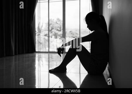 Silhouette einer depressiven jungen Frau in der Nähe des Fensters zu Hause, im Dunkeln. Häusliche Gewalt, Gewalt gegen Frauen Schwarzes und weißes Foto. Stockfoto