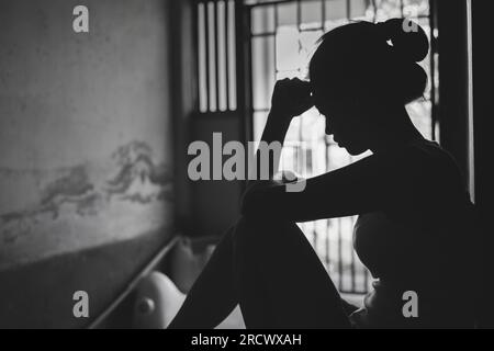 Silhouette einer depressiven jungen Frau in der Nähe des Fensters zu Hause, im Dunkeln. Häusliche Gewalt, Gewalt gegen Frauen Schwarzes und weißes Foto. Stockfoto