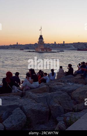 Die Menschen sitzen auf Felsen und genießen den Sonnenuntergang über dem Bosporusmeer und dem Maidens Tower von Uskudar, Istanbul, Türkei. Galata Tower und Kreuzfahrtschiff dahinter. Stockfoto