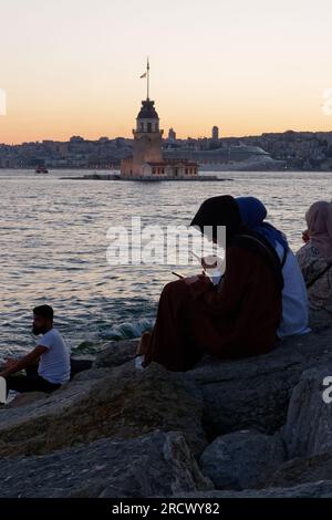 Die Menschen sitzen auf Felsen und genießen den Sonnenuntergang über dem Bosporusmeer und dem Maidens Tower von Uskudar, Istanbul, Türkei. Kreuzfahrtschiff hinten. Stockfoto