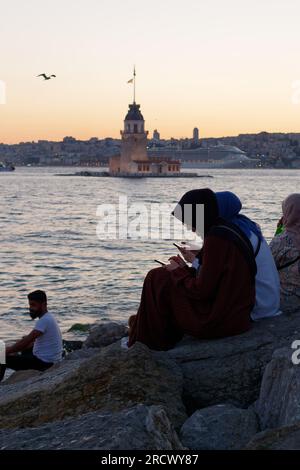 Die Menschen sitzen auf Felsen und genießen den Sonnenuntergang über dem Bosporusmeer und dem Maidens Tower von Uskudar, Istanbul, Türkei. Kreuzfahrtschiff hinten. Stockfoto